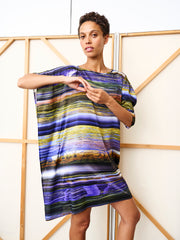 A person poses with one arm raised in front of a beige folding screen, wearing the Long-Sleeved Contra Dress by Zero + Maria Cornejo, featuring multicolor horizontal stripes in blue, purple, yellow, and green.