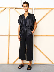 A person stands indoors against a wooden panel, hands in pockets, wearing Zero + Maria Cornejo's black Ruched Front Pant and a loose black satin top with slide sandals. Made in New York.