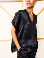 A woman stands against a light background in the black Fin Gaban Shirt by Zero + Maria Cornejo, styled with matching pants and one hand in her pocket.