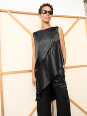 A person in sunglasses stands before a wooden geometric panel, wearing the black Heli Tunic by Zero + Maria Cornejo, styled with matching wide-leg pants. The elegant asymmetrical sleeveless tunic is made in New York.