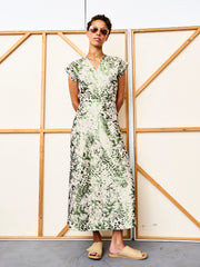 A person in sunglasses, beige sandals, and the Zero + Maria Cornejo Wrap Omi Dress—a light green and white patterned metallic jacquard with a wrap v-neck—stands with hands behind their back before a wooden-framed backdrop.