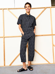 A person stands smiling with hands in their pockets, wearing the black Abi Pant by Zero + Maria Cornejo and black slide sandals, set against a light background with wooden framing.