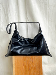A black, slouchy hobo-style Bartleby Objects Large Kestor Bag with an adjustable strap, resting on a wooden pedestal.