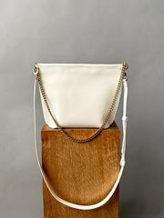 The Bartleby Objects Arden Bag, a white crossbody with gold chain and leather straps, is displayed on a textured wooden block against a plain gray background.