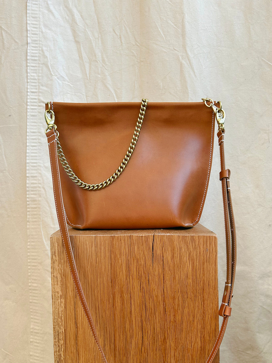 The Bartleby Objects Arden Bag, crafted from brown vachetta leather, featuring a gold chain and adjustable shoulder strap, is displayed on a wooden surface against a cream-colored fabric background.