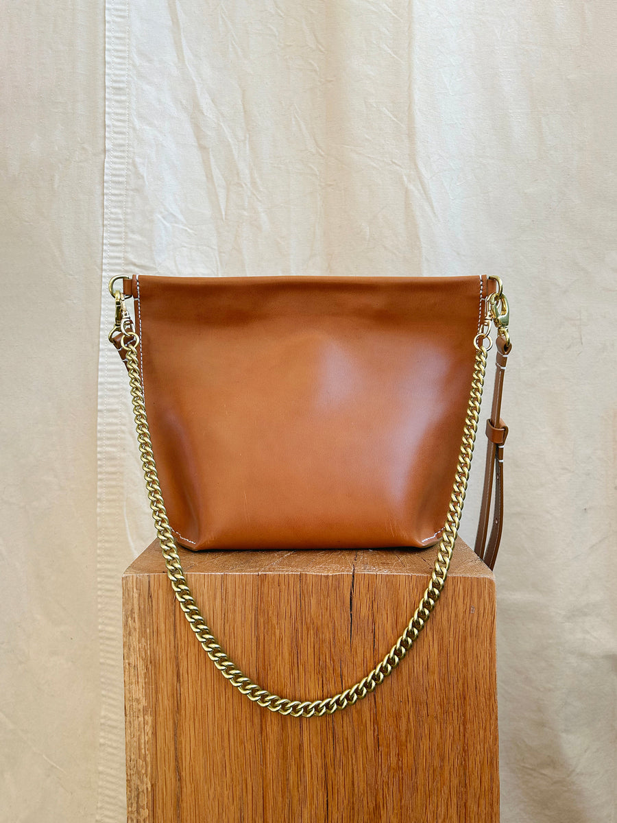 The Bartleby Objects Arden Bag, a chic brown vachetta leather handbag with a gold chain strap, is elegantly showcased on a wooden block. The light-colored fabric curtain background embodies the minimalist elegance for which Bartleby Objects is known.