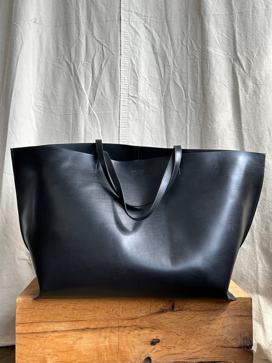 The Bartleby Objects Declan XL Tote, a handmade extra-large black leather bag from New York City, features a minimal design and rests on a wooden surface. Its sleek waxy vachetta leather perfectly complements the neutral-colored fabric curtain in the background.