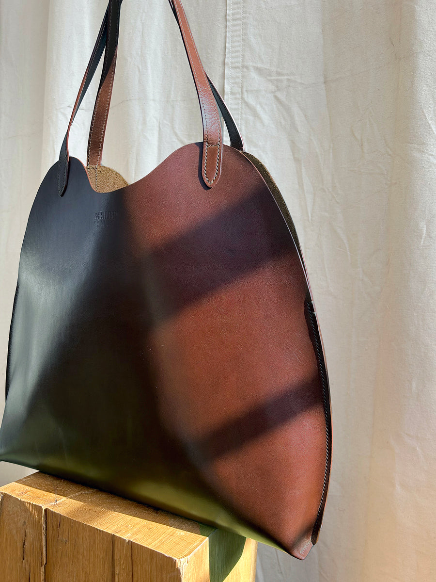 The Bartleby Objects Fawkes Tote, handmade from brown vegetable-tanned leather, rests on a wooden surface with sunlight casting shadows, set against a white fabric backdrop.