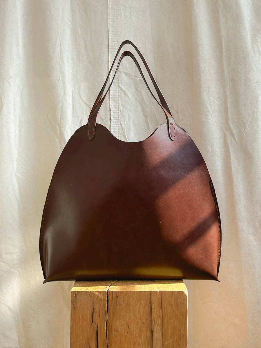 The Bartleby Objects Fawkes Tote, a large handmade bag with curved edges and two handles, rests on a wooden block against a sunlit neutral fabric backdrop.