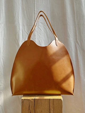 The Bartleby Objects Fawkes Tote, a handmade piece from Bartleby Objects, features a rounded shape and thin handles, crafted from brown vegetable-tanned leather. It is showcased on a wooden block against softly draped fabric in natural lighting.