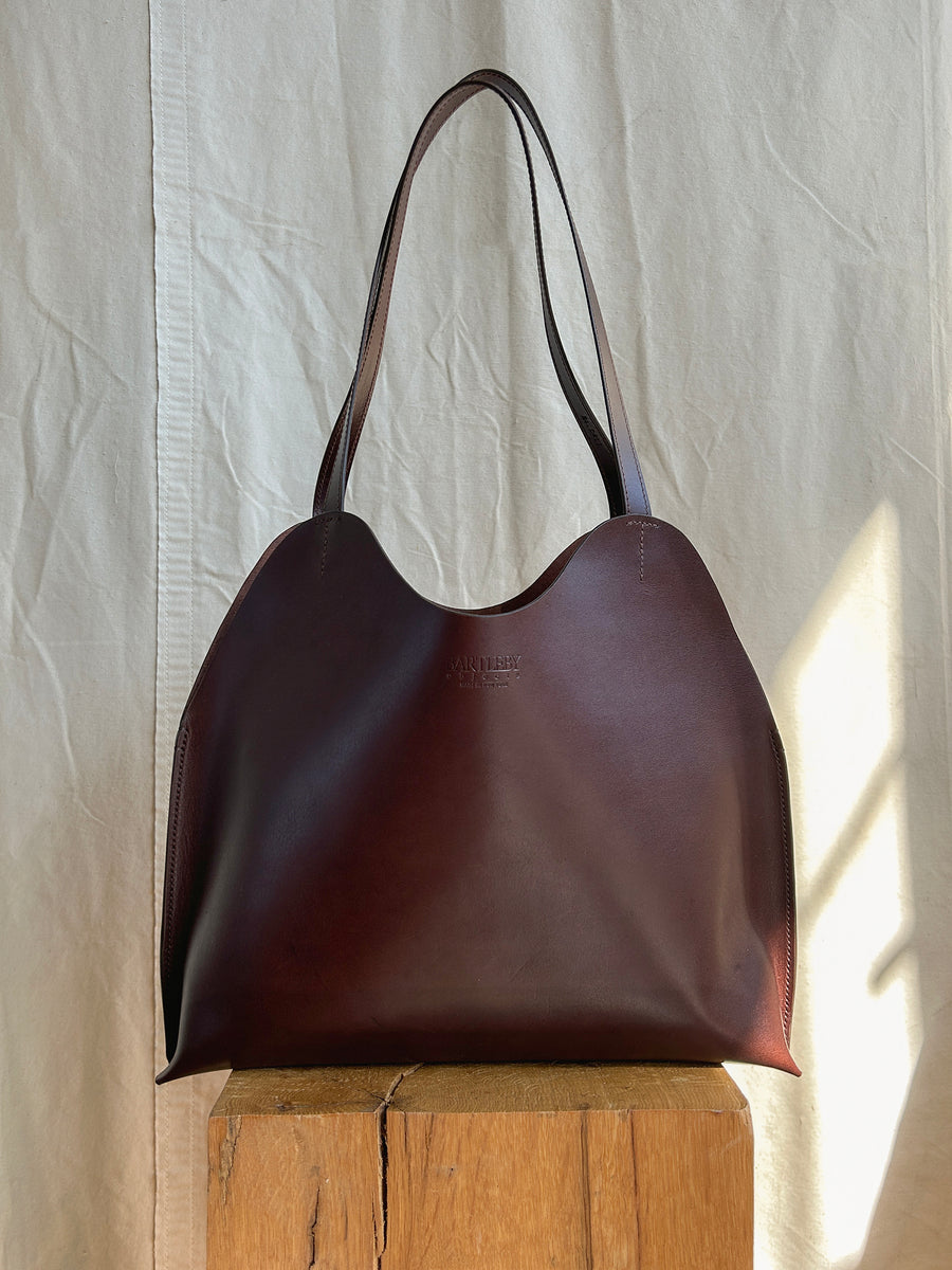 The Bartleby Objects Freja Tote, a large chocolate vegetable-tanned leather bag, handmade in New York City, sits on a wooden block against a cream textured fabric backdrop.