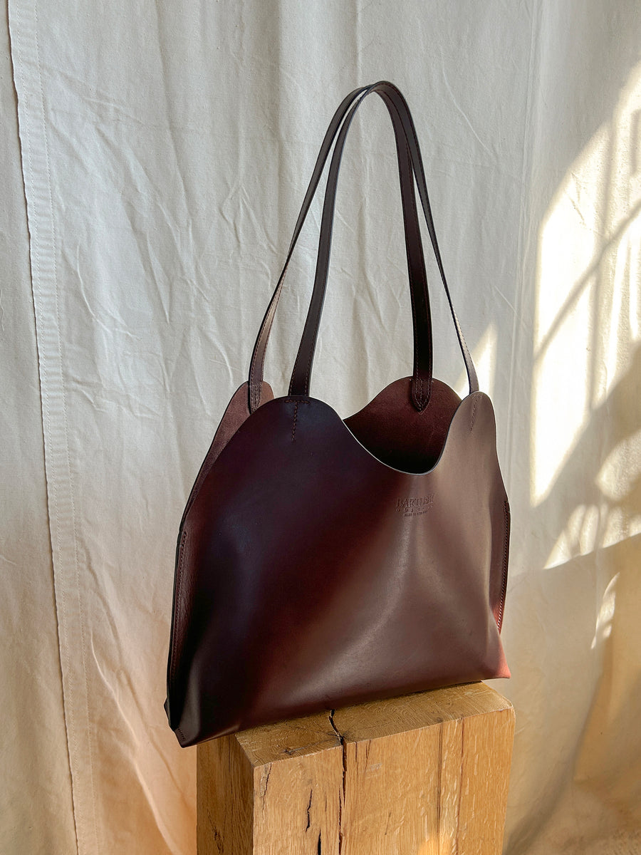 The Bartleby Objects Freja Tote, a dark brown vegetable-tanned leather bag with wavy edges and two handles, handmade in NYC, sits on a wooden block against light fabric as sunlight creates soft shadows.