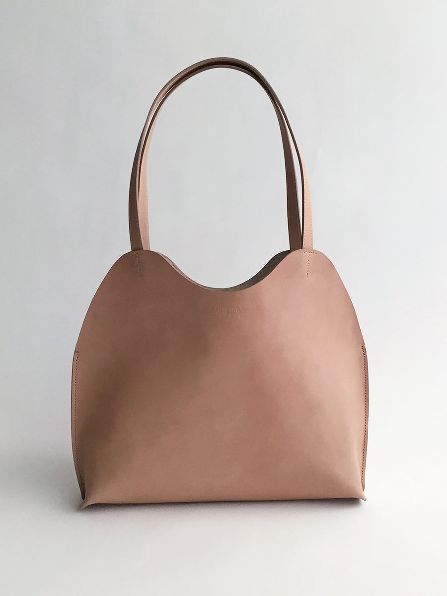 The Bartleby Objects Freja Tote, a light brown handbag crafted from vegetable-tanned leather, showcases a smooth surface and two straps. Set against a neutral background, this New York City handmade piece features a curved top design.