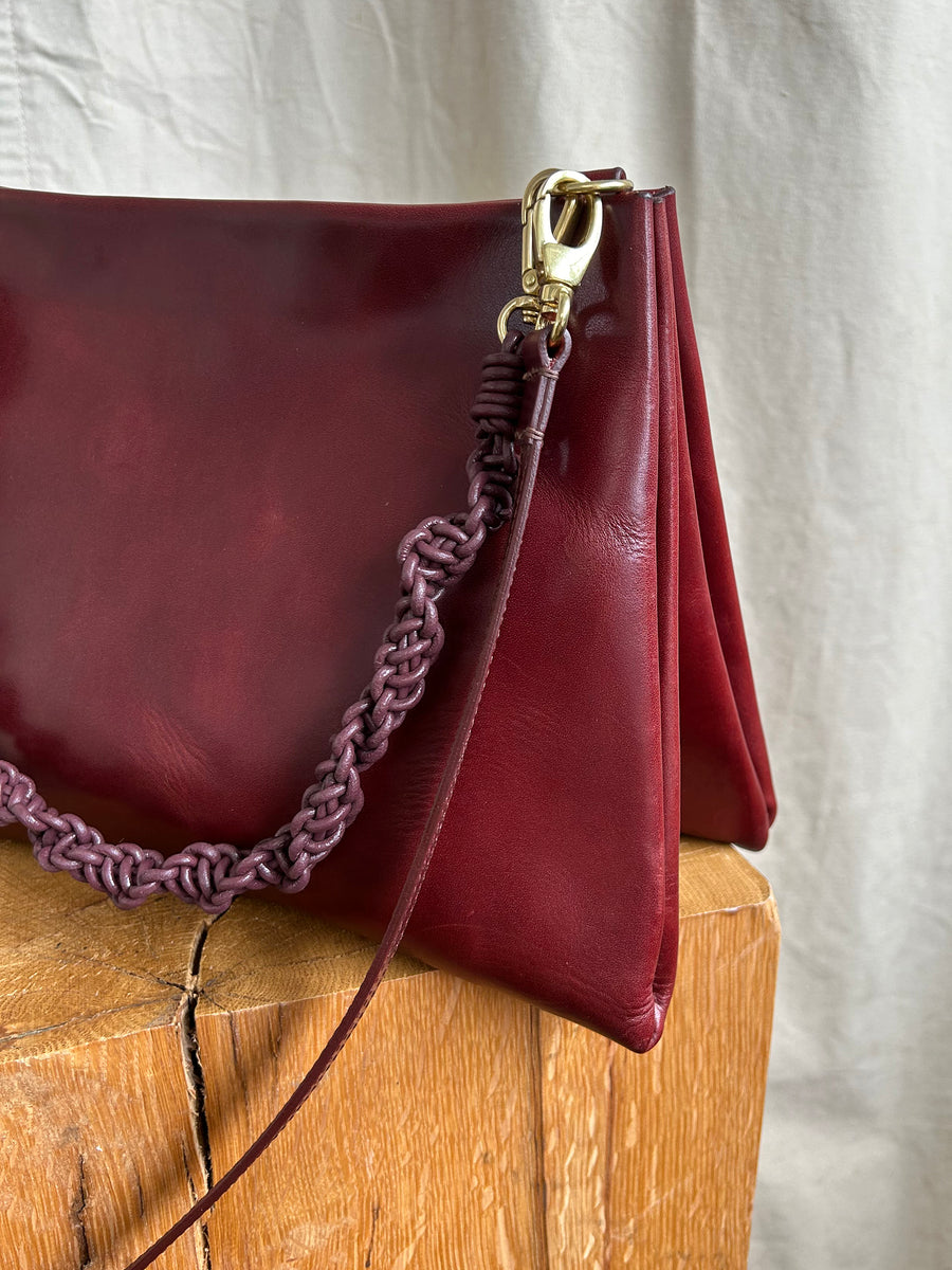 A close-up of the Bartleby Objects Glynne Bag, a deep red leather crossbody handbag with a braided strap, handmade in New York City. The bag showcases gold hardware and is set on a wooden surface against a light fabric backdrop.