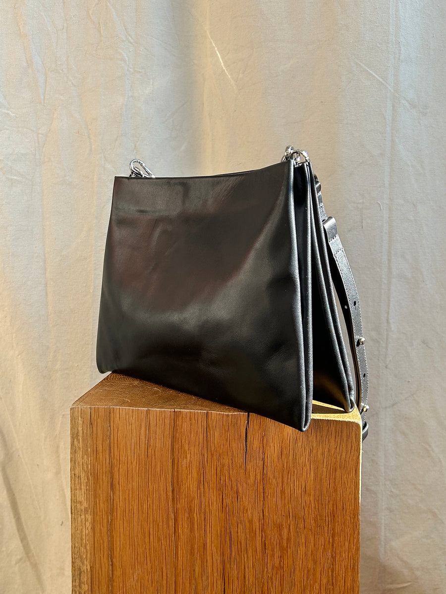 The Bartleby Objects Glynne Bag, a sleek black leather crossbody with adjustable straps, is elegantly displayed on a wooden pedestal against a textured beige background, proudly handcrafted in New York City.