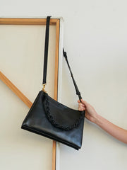 A hand holds the Bartleby Objects Glynne Bag—a black leather crossbody from Bartleby Objects featuring a ruched handle and long shoulder strap—set against a wooden frame and plain white wall.