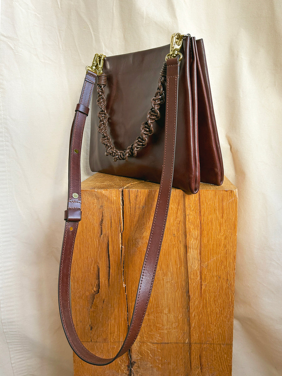 The Bartleby Objects Glynne Bag, a dark brown leather crossbody with gold clasp, ruched handle, and adjustable strap, handmade in New York by Bartleby Objects, is shown on a wooden block against a cream fabric backdrop.