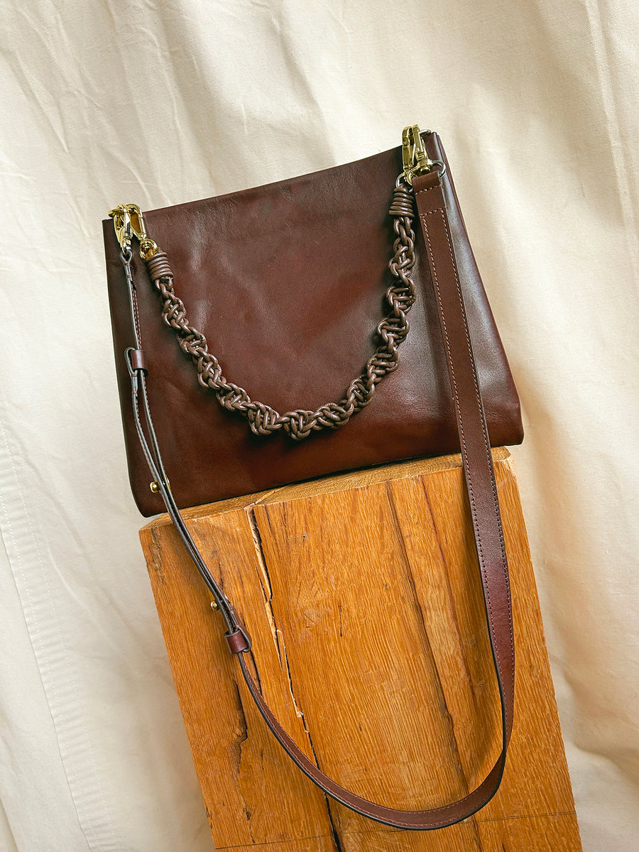 The Bartleby Objects Glynne Bag, a dark brown leather crossbody with twisted handle, adjustable strap, and double compartments, is shown upright on rustic wood with a beige fabric background. Handmade in New York by Bartleby Objects.