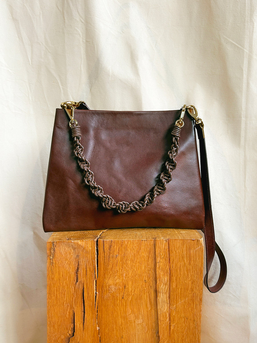 The Bartleby Objects Glynne Bag, a handmade dark brown leather crossbody with a braided handle and gold hardware by Bartleby Objects, rests on wood against a cream fabric backdrop.