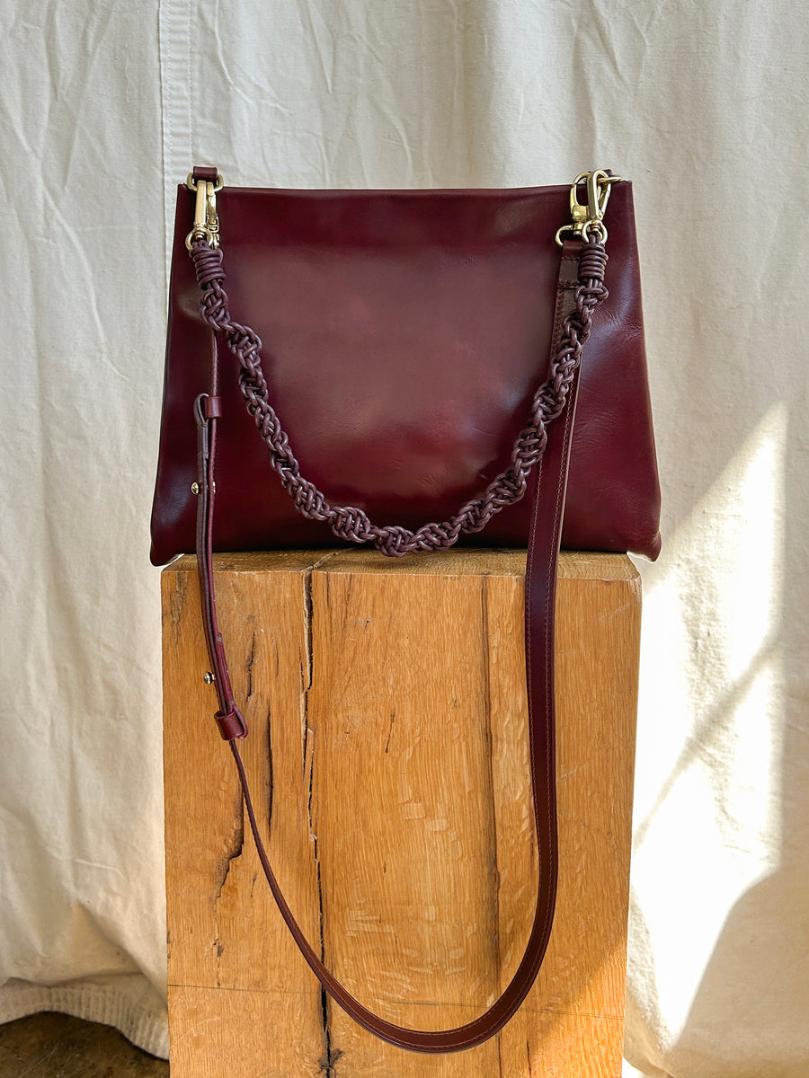 The Bartleby Objects Glynne Bag, a dark brown leather handbag with a braided leather strap and gold buckles, is displayed on a wooden block against a beige fabric backdrop. Handmade in New York City, natural light highlights the bag's smooth texture.