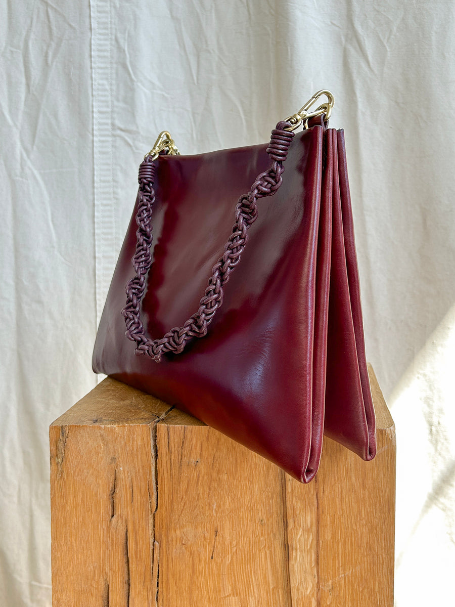 The Bartleby Objects Glynne Bag, a burgundy leather crossbody handbag featuring a braided chain and leather strap, is elegantly displayed on a wooden block against a light fabric backdrop, meticulously handmade in New York City.