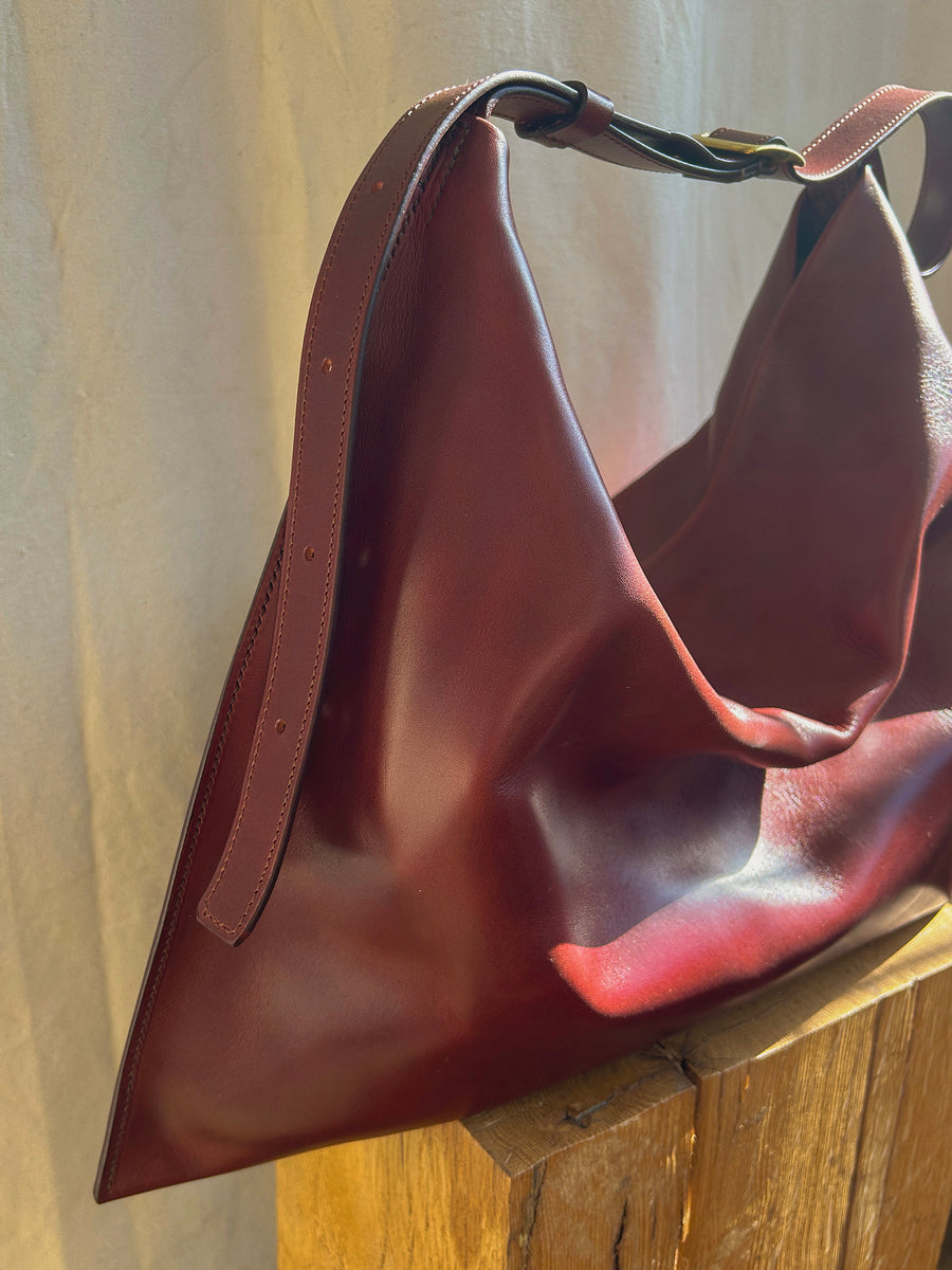 The Bartleby Objects Medium Kestor Bag, made from vegetable-tanned leather and crafted in New York City, rests on a wooden surface with its soft, slouchy shape. The sunlight accentuates the smooth texture and rich burgundy hue of the leather.