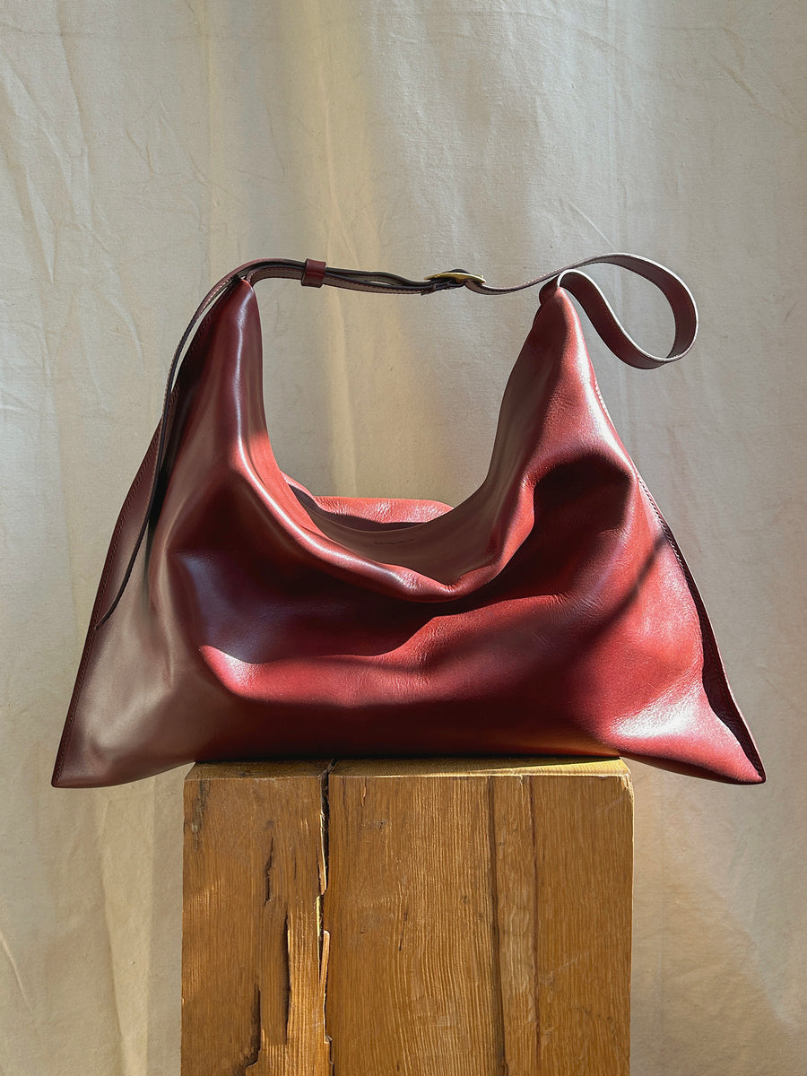 A medium-sized deep red slouchy rectangular bag, known as the Bartleby Objects Medium Kestor Bag, made of vegetable tanned leather hangs gracefully over a rustic wooden stool. The backdrop is a soft, neutral fabric, emphasizing the bag's rich color and texture. This exquisite piece by Bartleby Objects is handmade in New York City.