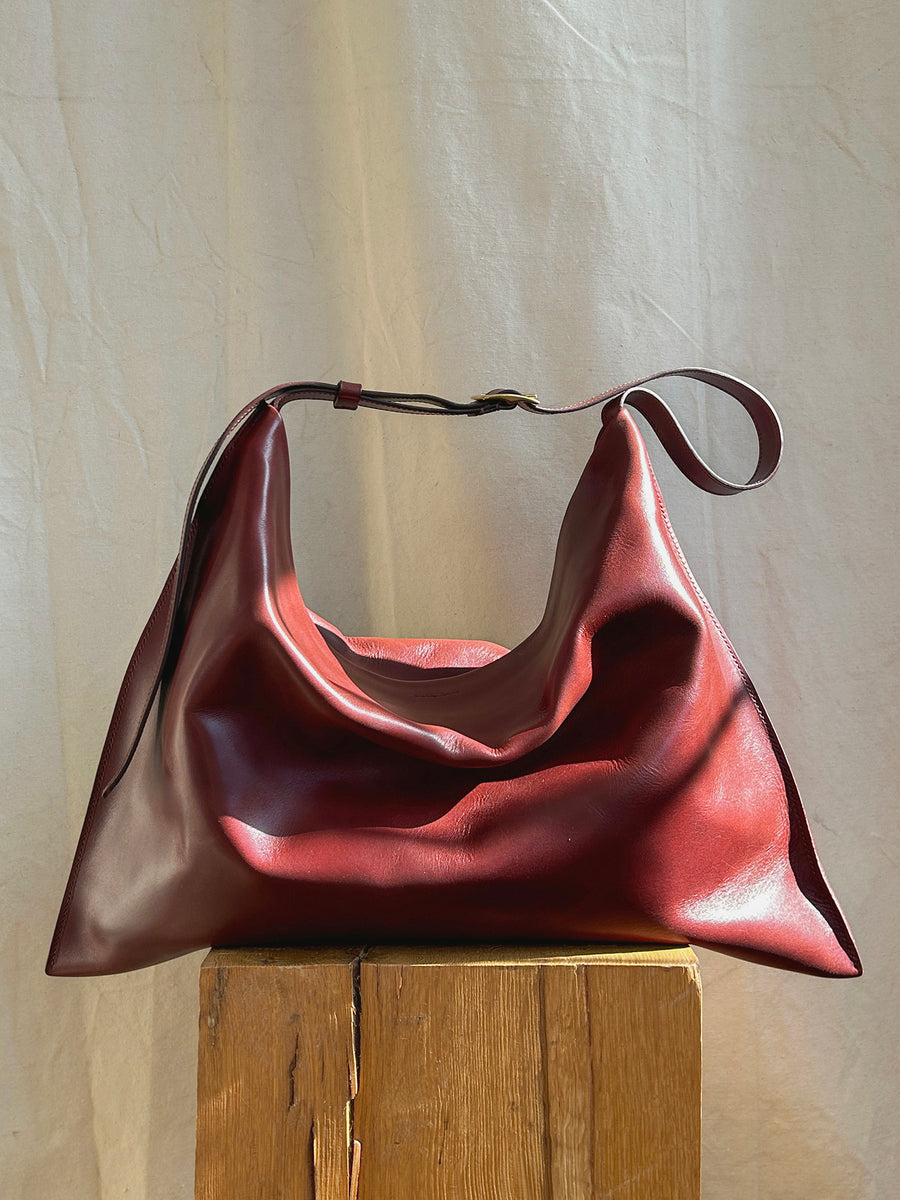 A medium Kestor Bag by Bartleby Objects, crafted from burgundy vegetable-tanned leather, rests on a wooden stool against a light-colored fabric backdrop. This handmade-in-New York City piece features a slouchy rectangular design with a single adjustable strap draped casually over the top.