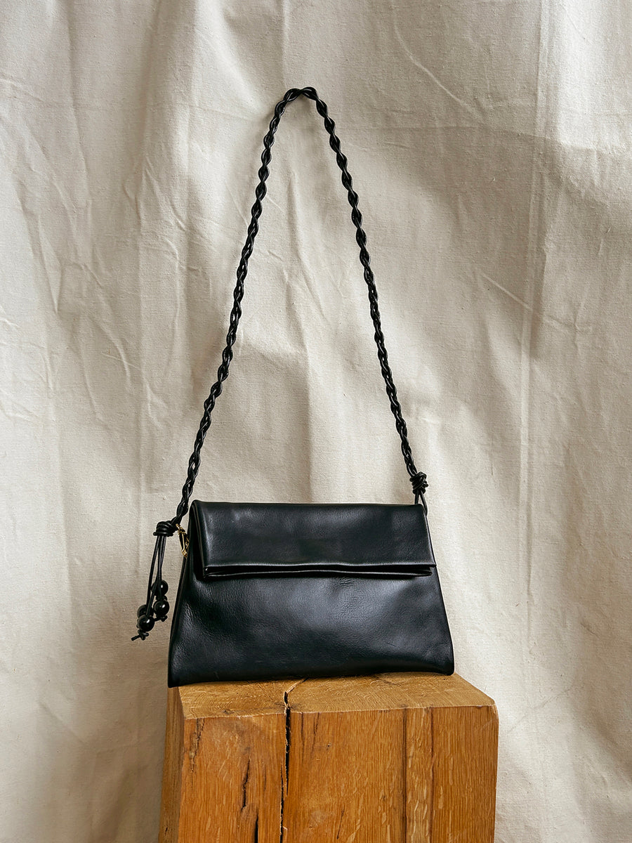 The Bartleby Objects Small Verani Crossbody, crafted from black vachetta leather with a braided strap, is displayed on a wooden block against a wrinkled beige fabric backdrop.