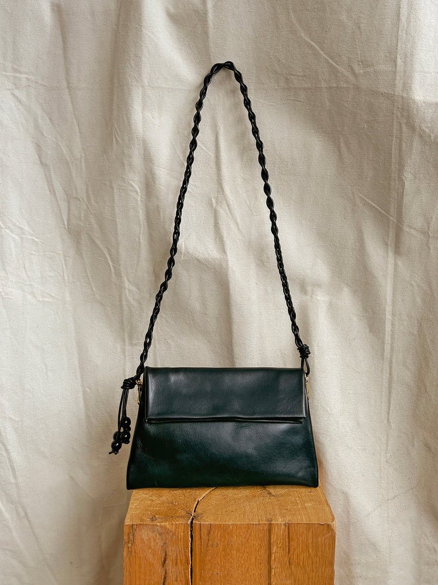The Bartleby Objects Small Verani Crossbody, a black vachetta leather handbag with a braided strap, is displayed on a wooden block against a light textured fabric background.