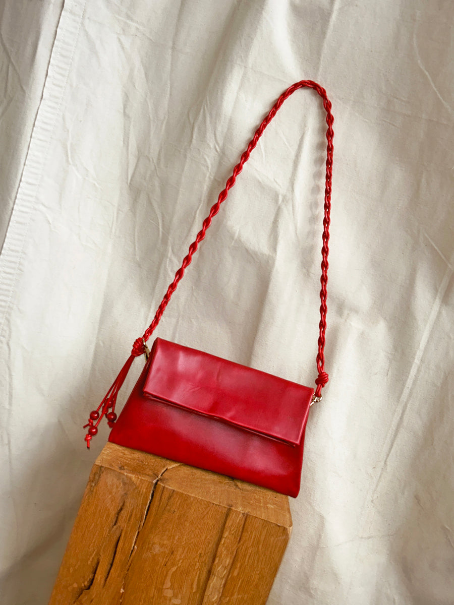 The Bartleby Objects Small Verani Crossbody, a handmade red bag with a braided strap by Bartleby Objects in NYC, is shown on a wooden block against a white fabric background.