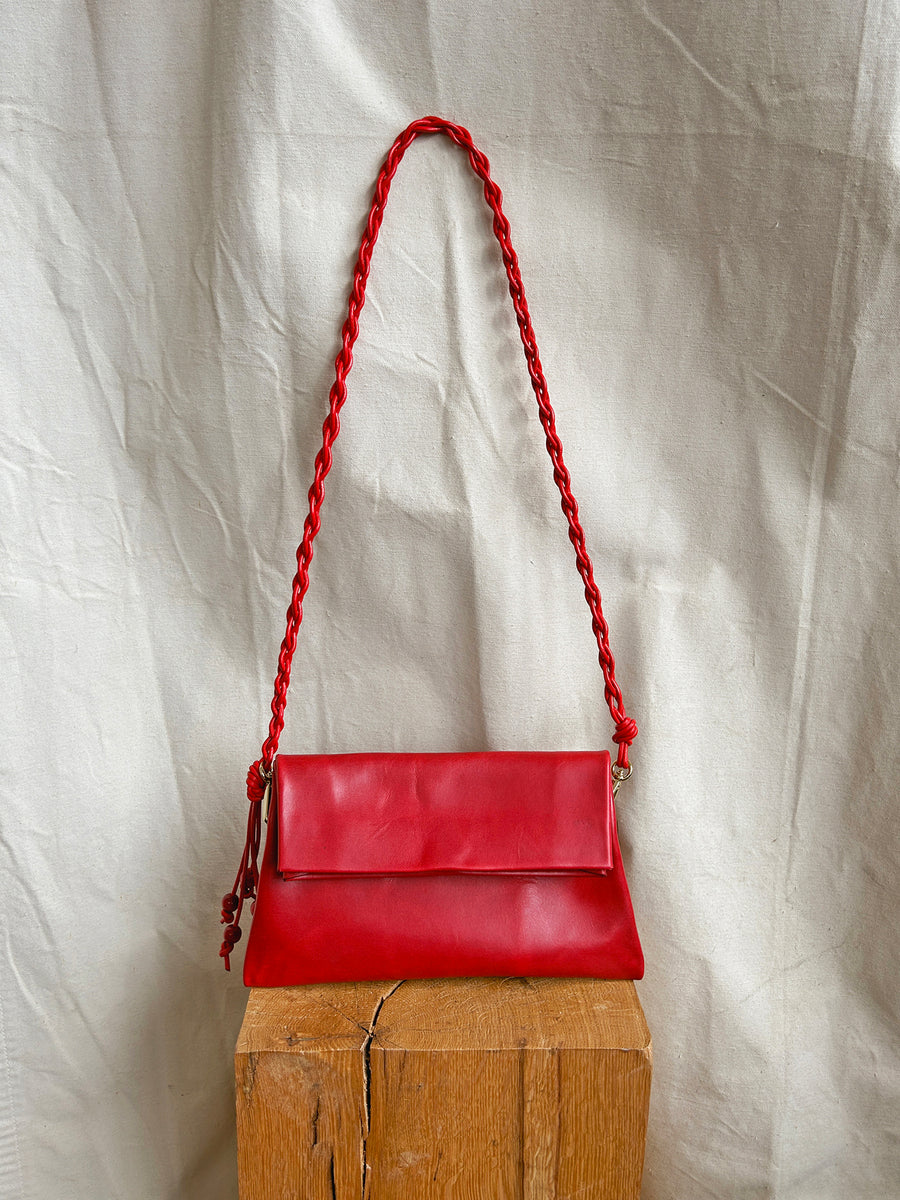 The Bartleby Objects Small Verani Crossbody, a red vachetta leather bag with a braided strap, is handmade in New York City and shown on a wooden block against a cream-colored fabric backdrop.