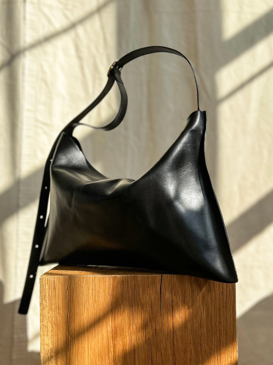 The Bartleby Objects Small Kestor Bag, a black, rectangular handbag made of vegetable-tanned leather, is showcased on a wooden surface. Sunlight highlights its smooth texture and adjustable strap, reflecting its expert craftsmanship from New York City.