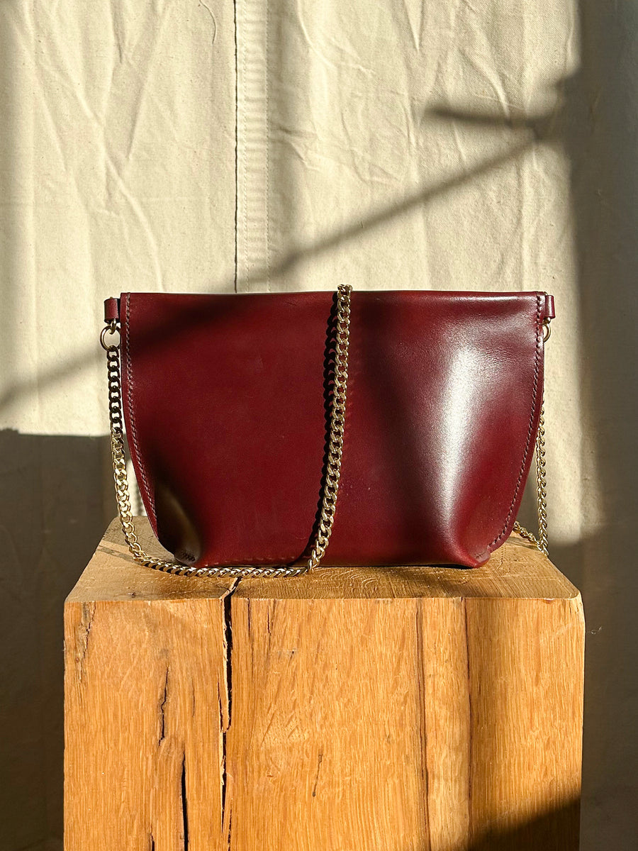 The Bartleby Objects Stowe Chain Bag, a handmade piece crafted from burgundy leather and featuring a solid brass chain strap, is displayed on a wooden block. The scene is set against a textured fabric background, with shadows cast by the sunlight.