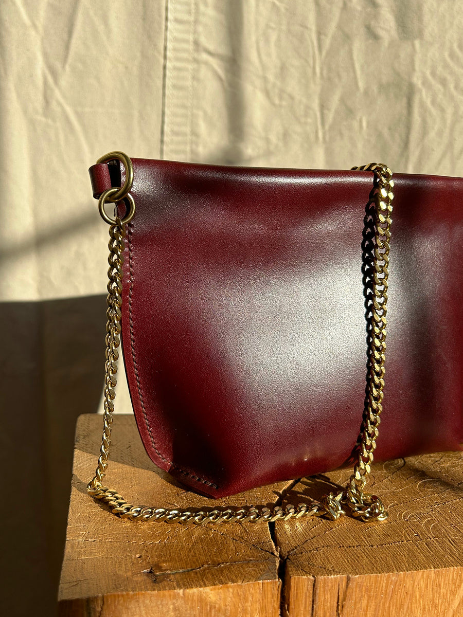 The Bartleby Objects Stowe Chain Bag, crafted from burgundy leather and featuring a solid brass chain strap, rests on a wooden surface. The backdrop showcases light-colored fabric with shadows playing across it, enhancing the sophistication of the vegetable-tanned leather.