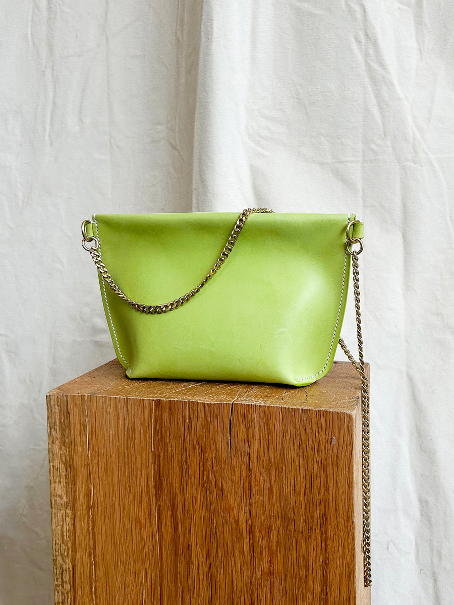 The Bartleby Objects Stowe Chain Bag, featuring a sleek design made from vegetable-tanned leather, is displayed on a wooden pedestal against a white fabric backdrop. This lime green handbag, handmade in New York City, includes a solid brass chain strap and a stylish fold-over flap.