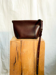Dark brown vegetable-tanned leather Bartleby Objects Stowe Leather Strap Bag with an adjustable strap resting on a wooden block.