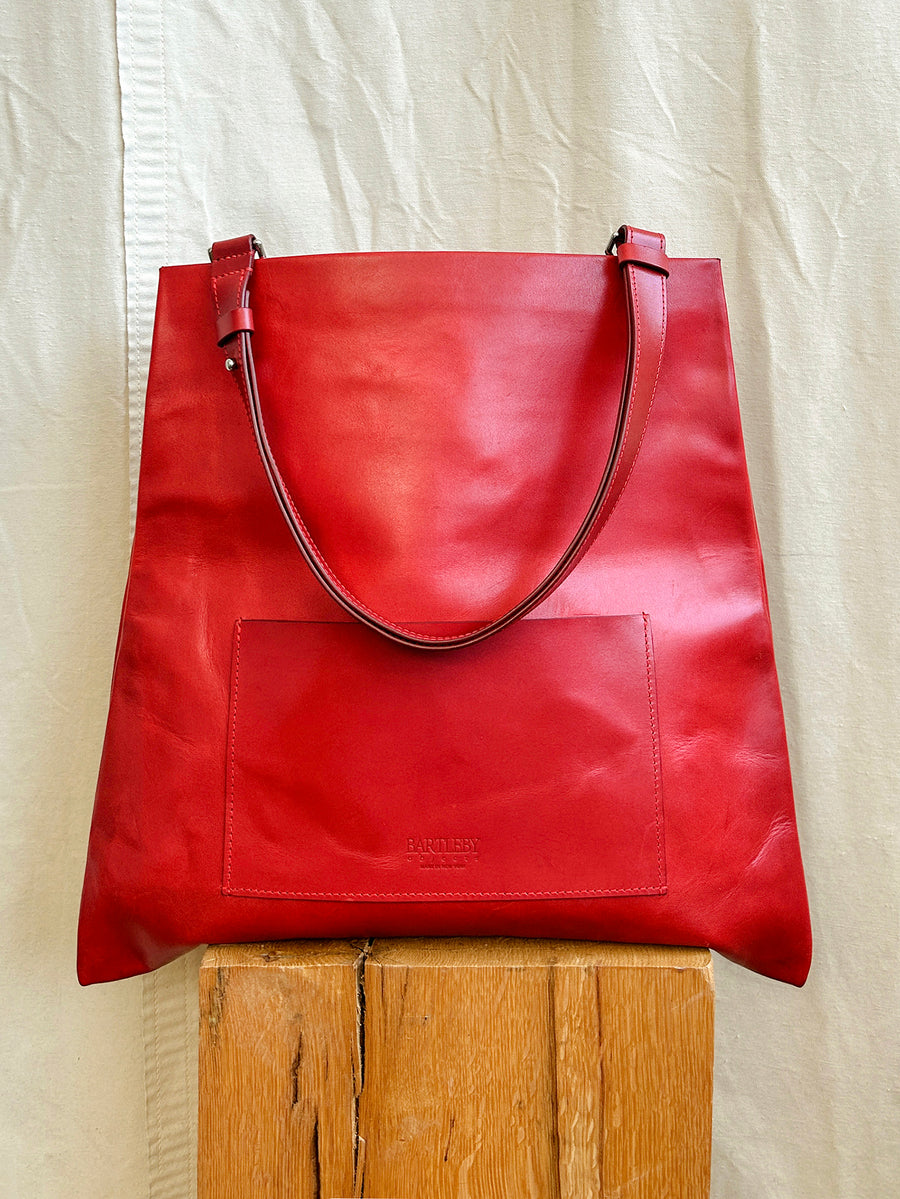 The Bartleby Objects Verani Large Convertible Clutch by Bartleby Objects is a large, bright red clutch handmade in NYC from waxy vachetta leather, shown upright on a wooden block against a cream-colored fabric background.