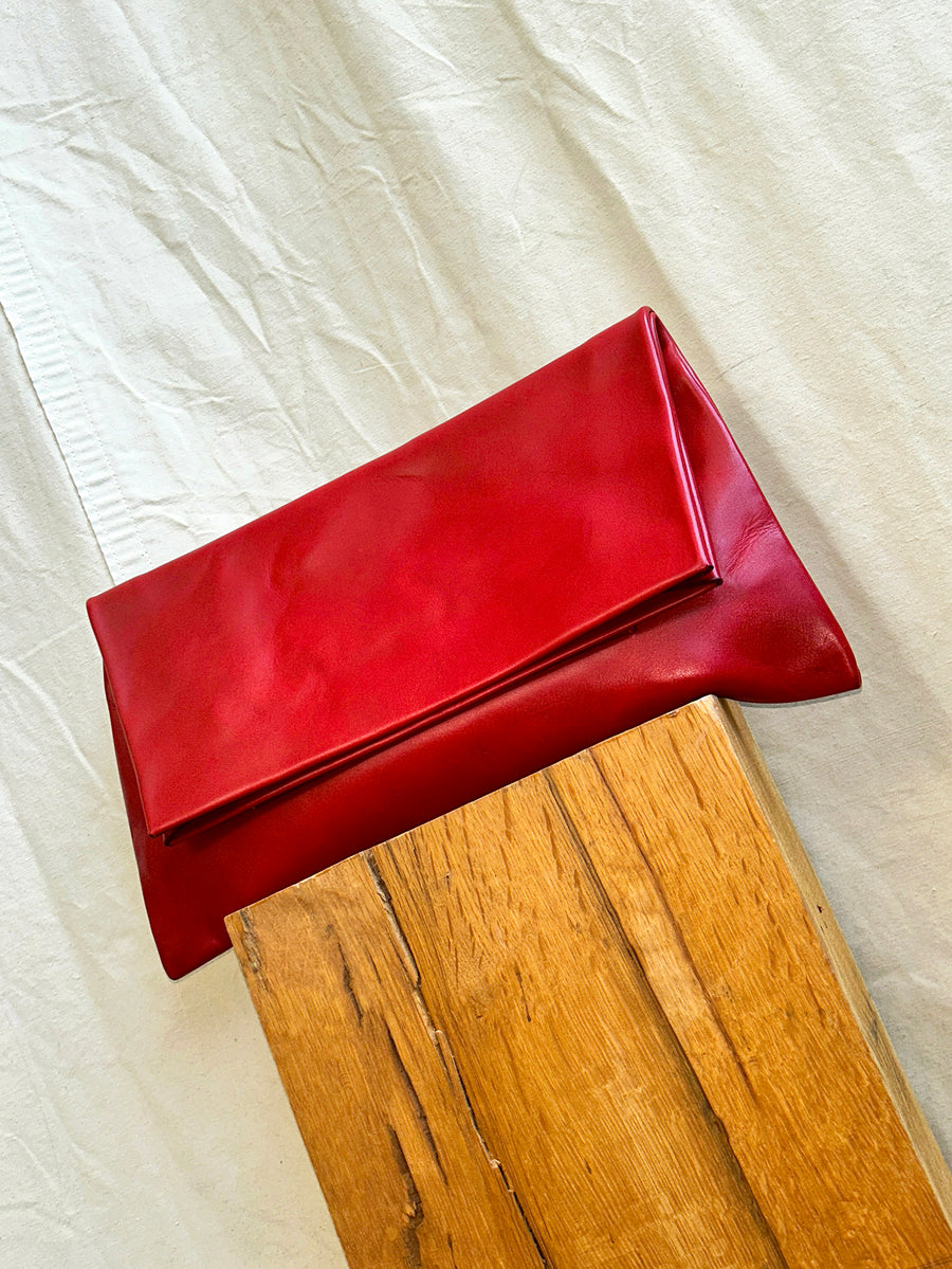 The Bartleby Objects Verani Large Convertible Clutch, a vibrant red bag crafted by Bartleby Objects in New York City from waxy vachetta leather, sits on wood with cream fabric behind it.