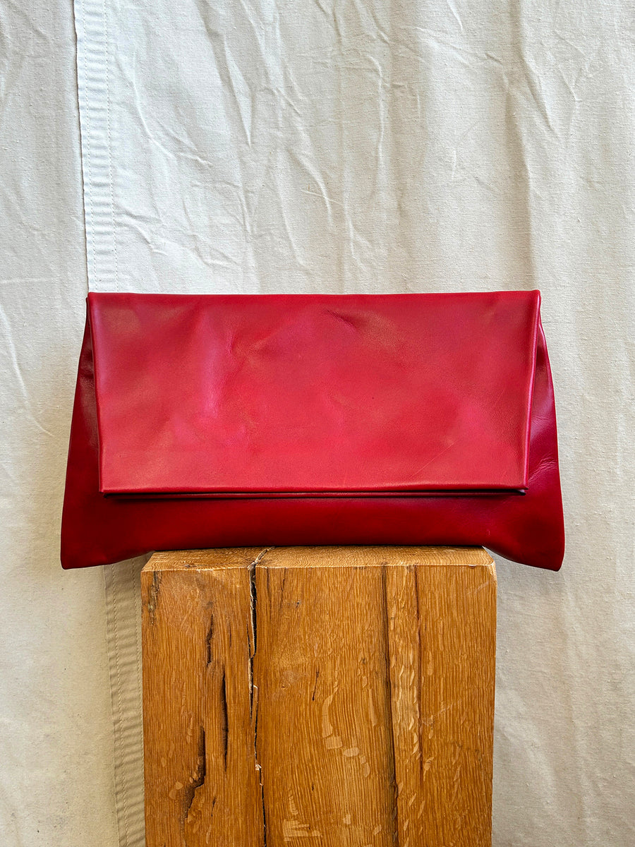 The Bartleby Objects Verani Large Convertible Clutch in red waxy vachetta leather stands upright on a wooden block, set against a textured beige fabric backdrop.