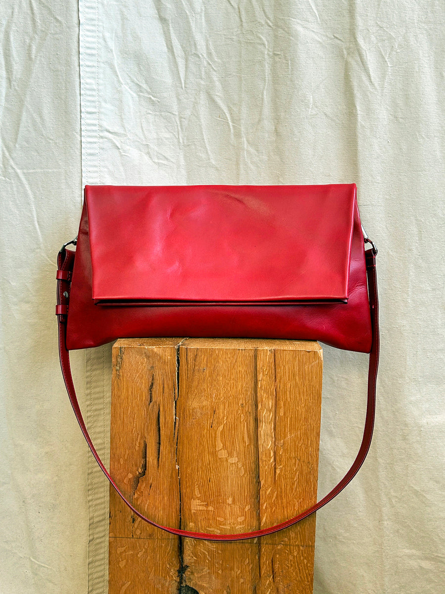 The Bartleby Objects Verani Large Convertible Clutch by Bartleby Objects, crafted from waxy vachetta leather in red and handmade in New York City, hangs on a wooden block against a cream-colored fabric backdrop.