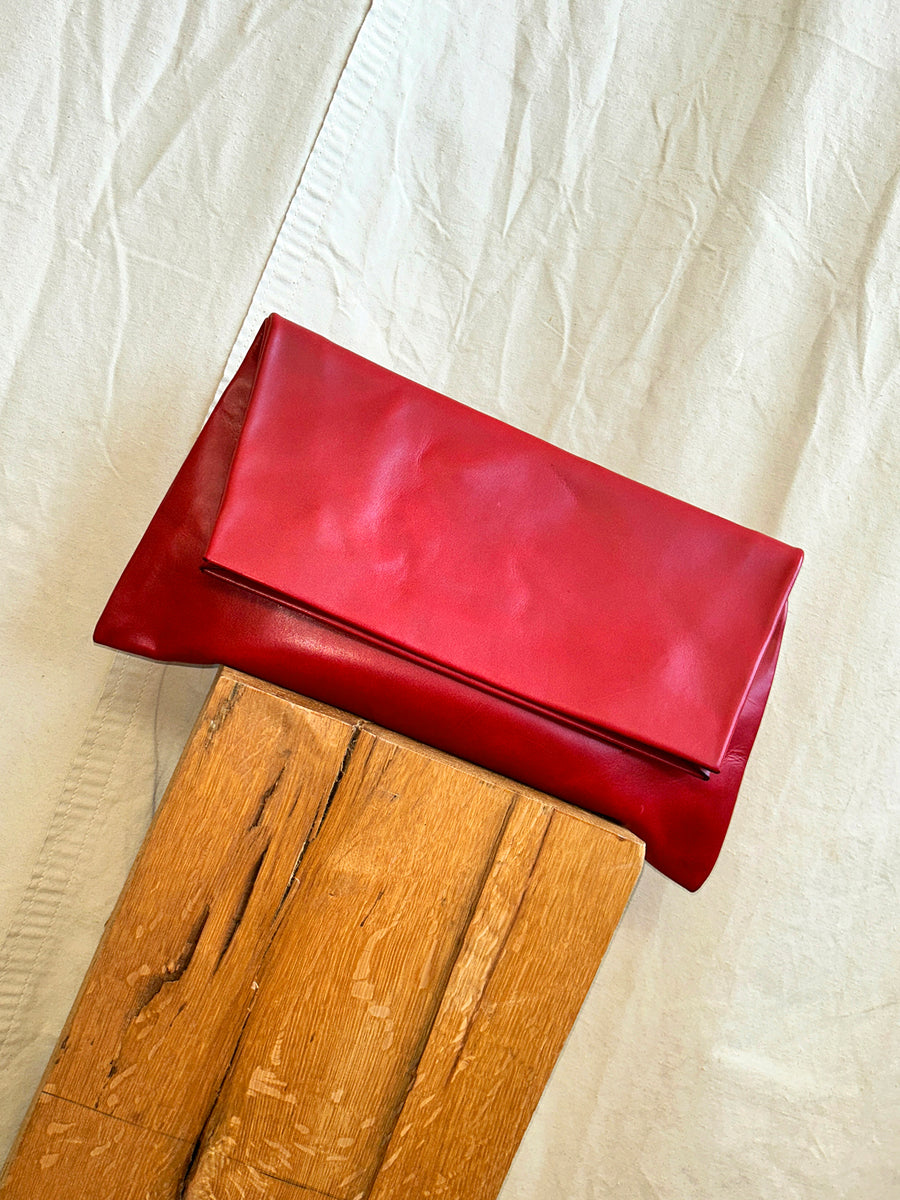 The Bartleby Objects Verani Large Convertible Clutch by Bartleby Objects, crafted from red waxy vachetta leather, sits angled on a wooden surface against a cream textured fabric backdrop.