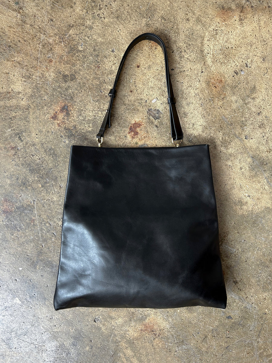 The Bartleby Objects Verani Large Convertible Clutch, a black leather bag by Bartleby Objects with a single shoulder strap, handmade in NYC, rests on a textured brown concrete floor.