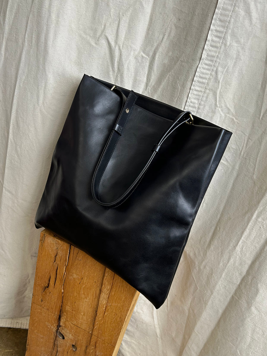 The Bartleby Objects Verani Large Convertible Clutch by Bartleby Objects, handmade in NYC from waxy vachetta leather with thin shoulder straps, is displayed on a wooden block against a beige fabric background.