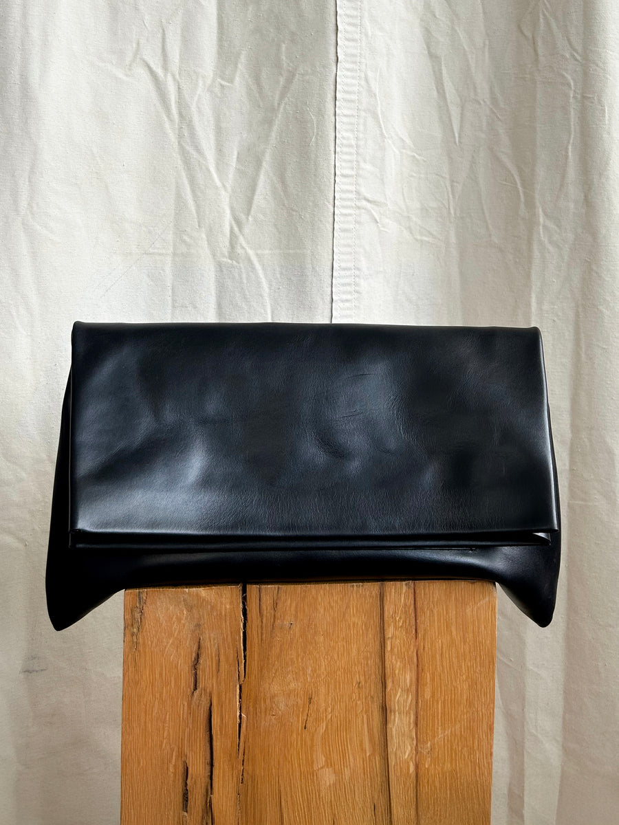 The Bartleby Objects Verani Large Convertible Clutch by Bartleby Objects, crafted from waxy vachetta leather in black and handmade in New York City, rests on a wooden block before a cream, wrinkled fabric backdrop.
