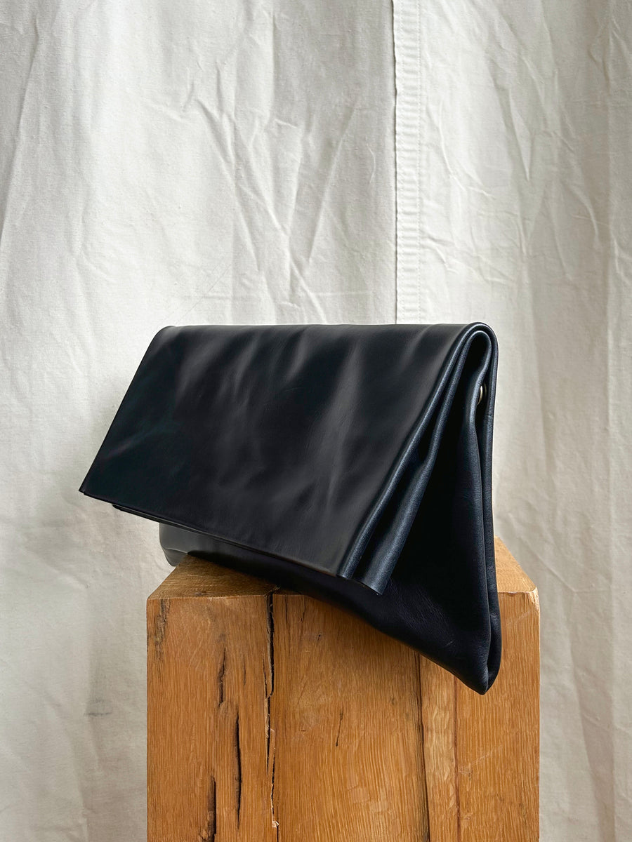 The Bartleby Objects Verani Large Convertible Clutch by Bartleby Objects, crafted from black waxy vachetta leather, features a triangular fold-over design and stands on a rustic wooden block against wrinkled white fabric, blending style with versatility.