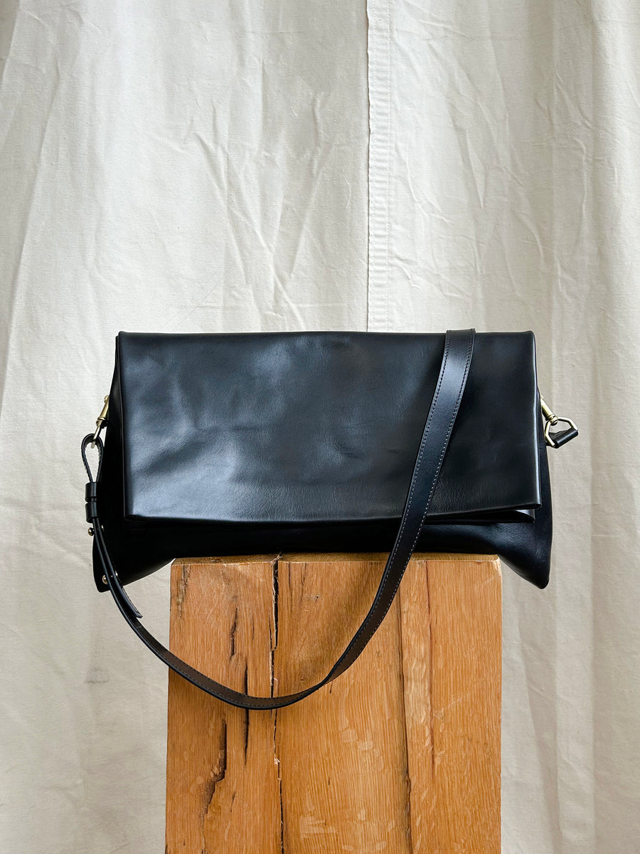 The Bartleby Objects Verani Large Convertible Clutch by Bartleby Objects, a black waxy vachetta leather shoulder bag with strap and flap closure, handmade in New York City, is shown on a light wooden block against a neutral fabric backdrop.