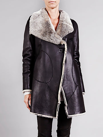 A person wears the Zero + Maria Cornejo Shearling Abbey Coat Archive Sample, featuring a long black design with an elegant draped collar and wide fluffy grey trim. With its asymmetrical front closure and rounded stitching details, the individual stands against a plain grey background.