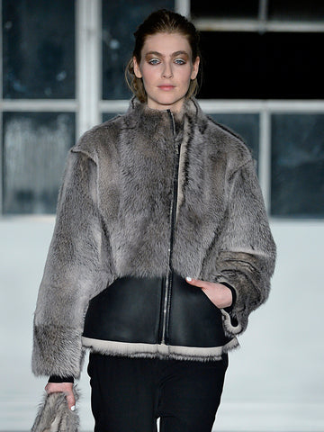 A model wearing a Zero + Maria Cornejo Shearling Sarki Jacket strides down the runway in black pants, posing confidently with one hand in her pocket. Large windows illuminate her lambskin ensemble against the spacious backdrop.