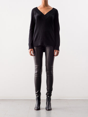 A person stands against a plain white background, wearing a black V-neck sweater, Zero + Maria Cornejo Leather Twist Seam Legging in black with an elastic waistband, and black ankle boots. Their arms are relaxed at their sides.